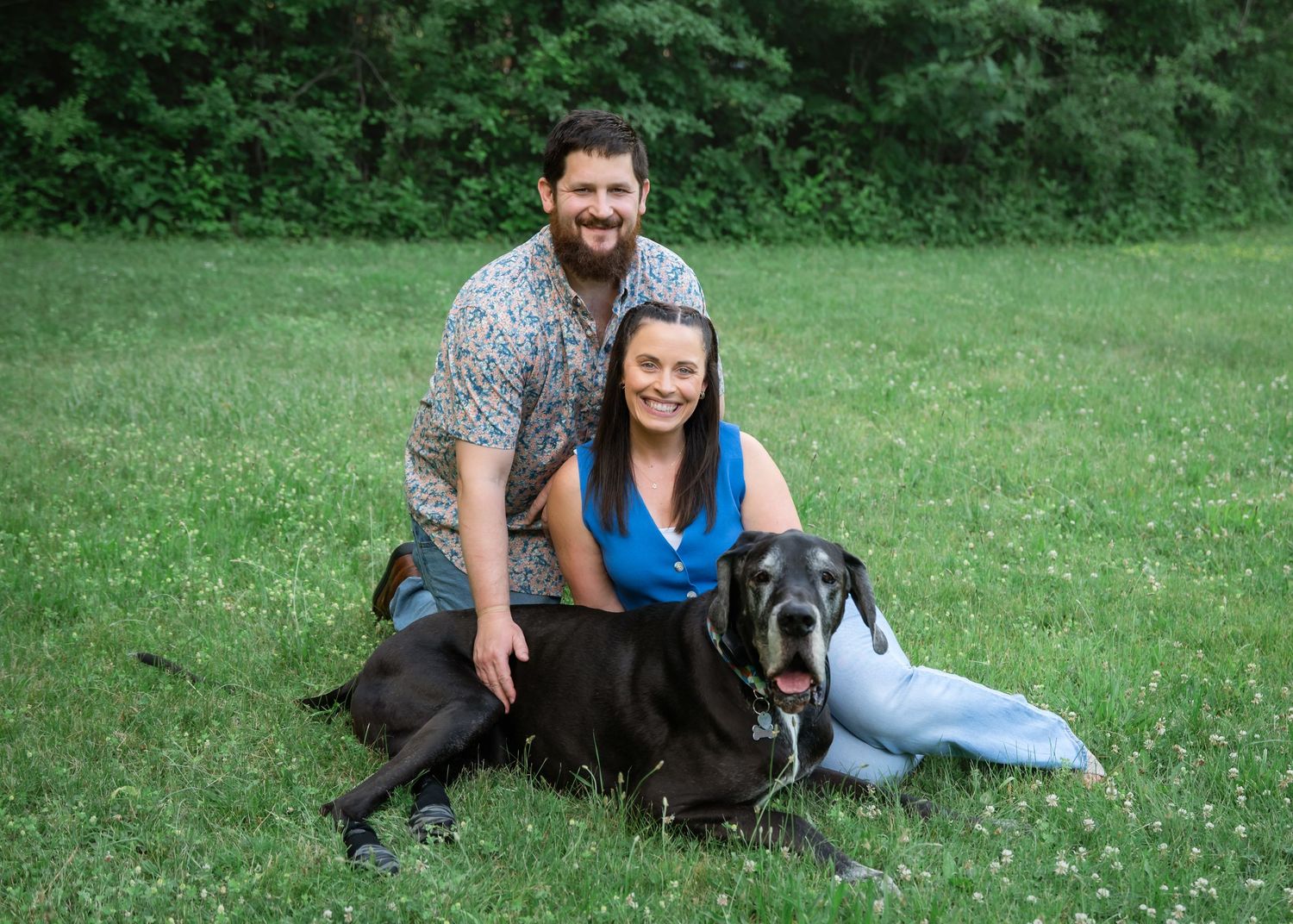 Couple smiling with a large black Great Dane dog on a green grassy lawn surrounded by trees.