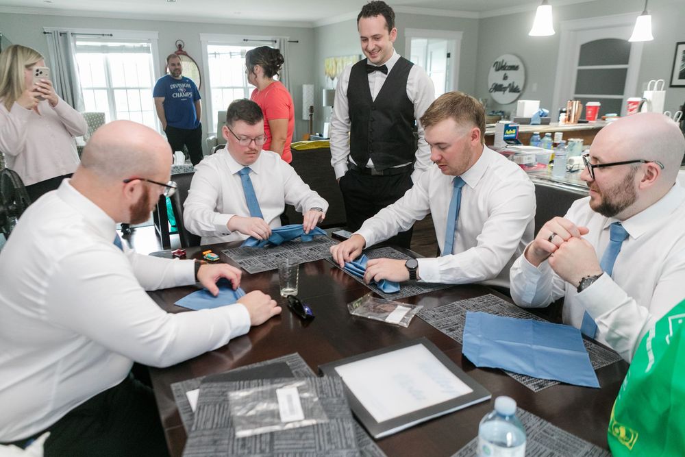 Groomsmen in white shirts and blue ties gather around a table preparing wedding accessories before the ceremony.