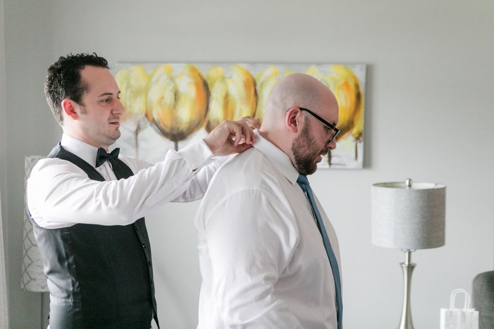 A groomsman in a black vest and bow tie helps a bald groom in a white shirt adjust his collar before the wedding.