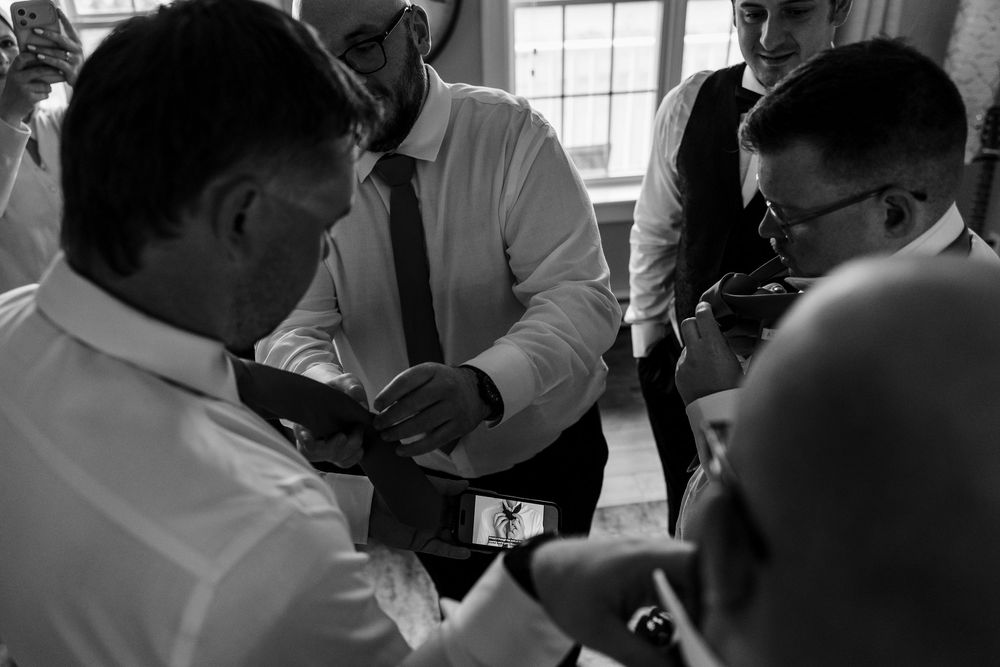 Black and white photo of groomsmen gathered together adjusting ties before a wedding ceremony.