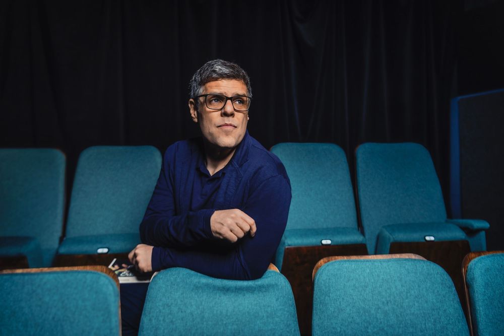 Man with glasses in blue sweater sits among teal theater seats, arms crossed, holding a phone.