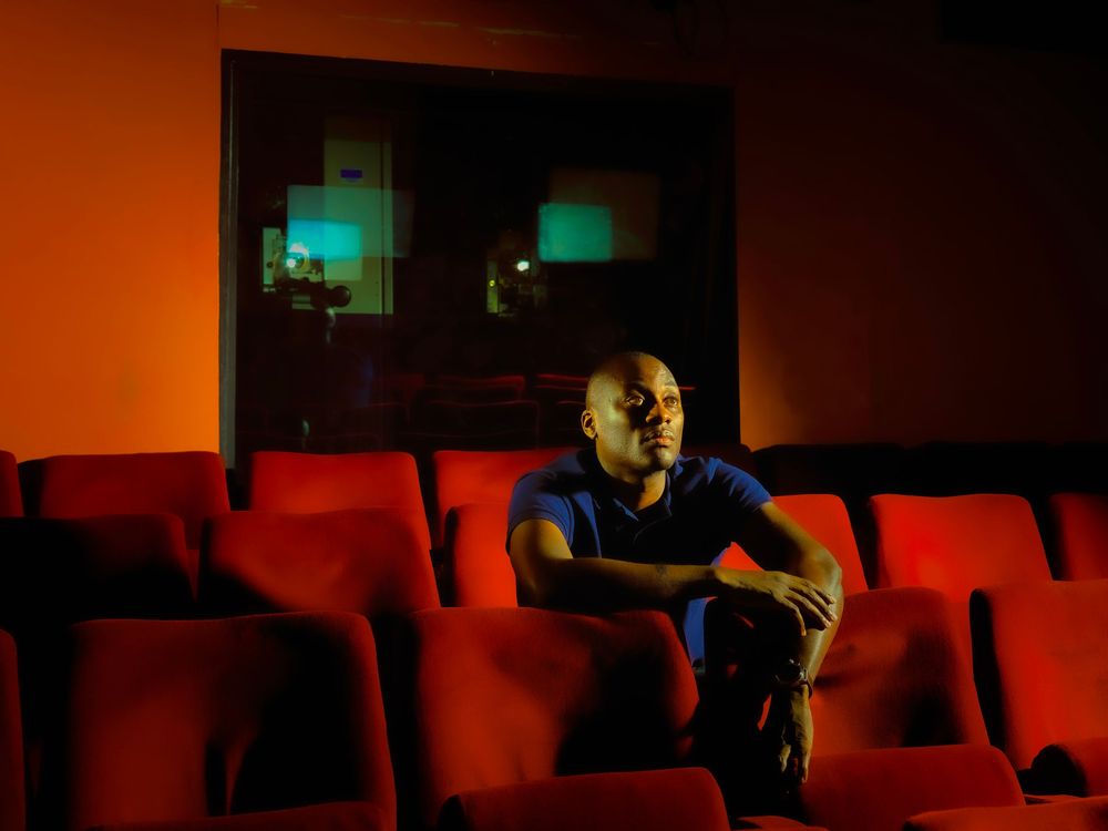 A man sits alone in red theater seats under warm orange lighting in an empty cinema.