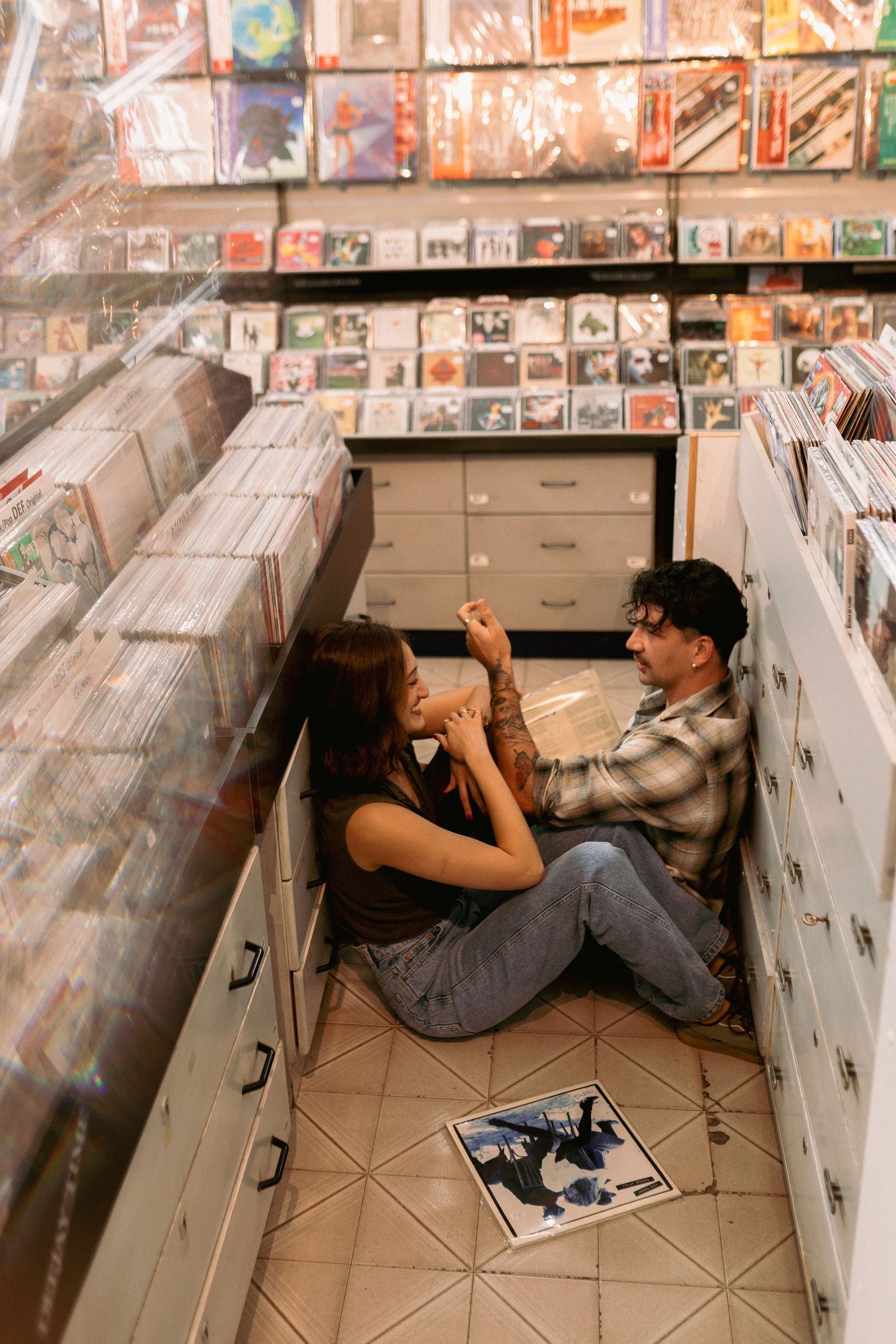 Couple sitting on floor of record store surrounded by vinyl albums and CDs, sharing a music discovery moment.