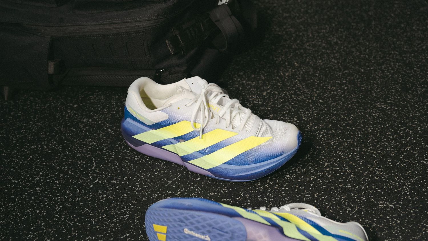 Adidas tennis shoes in white, yellow, and blue colorway placed on dark gym floor near a bag.