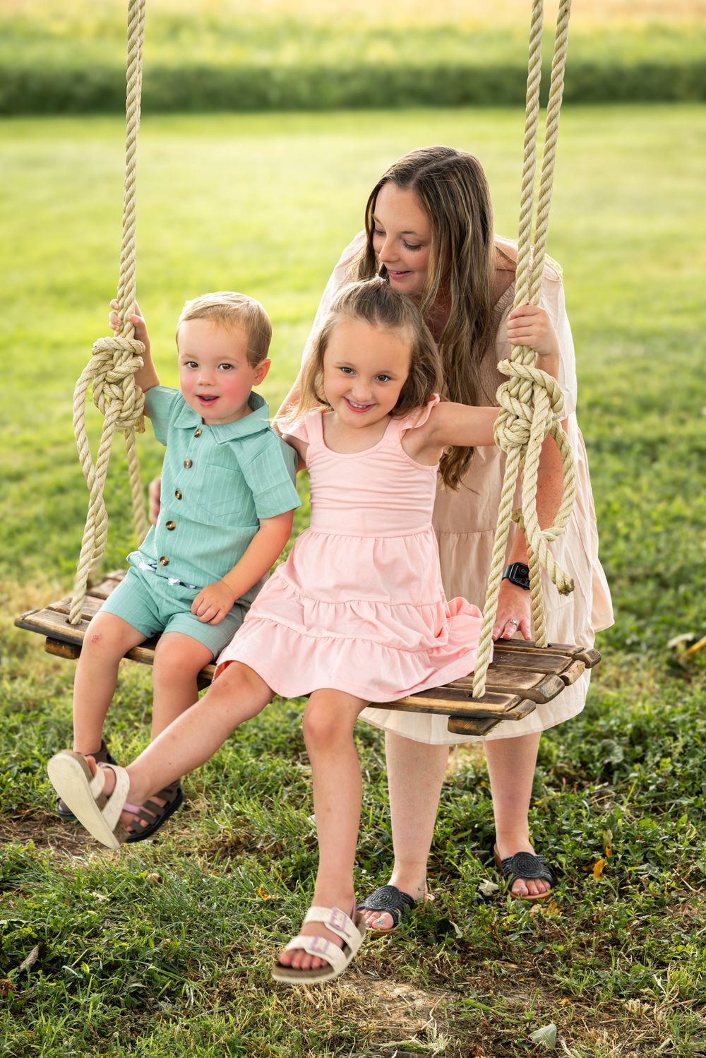 A mother and two young children sitting together on a wooden rope swing outdoors on a sunny day.