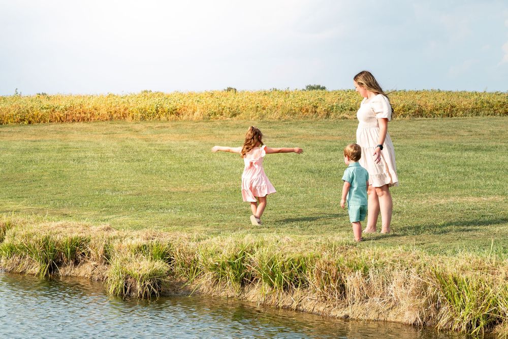 Pregnant mother with two young children playing near a pond on a sunny green meadow.