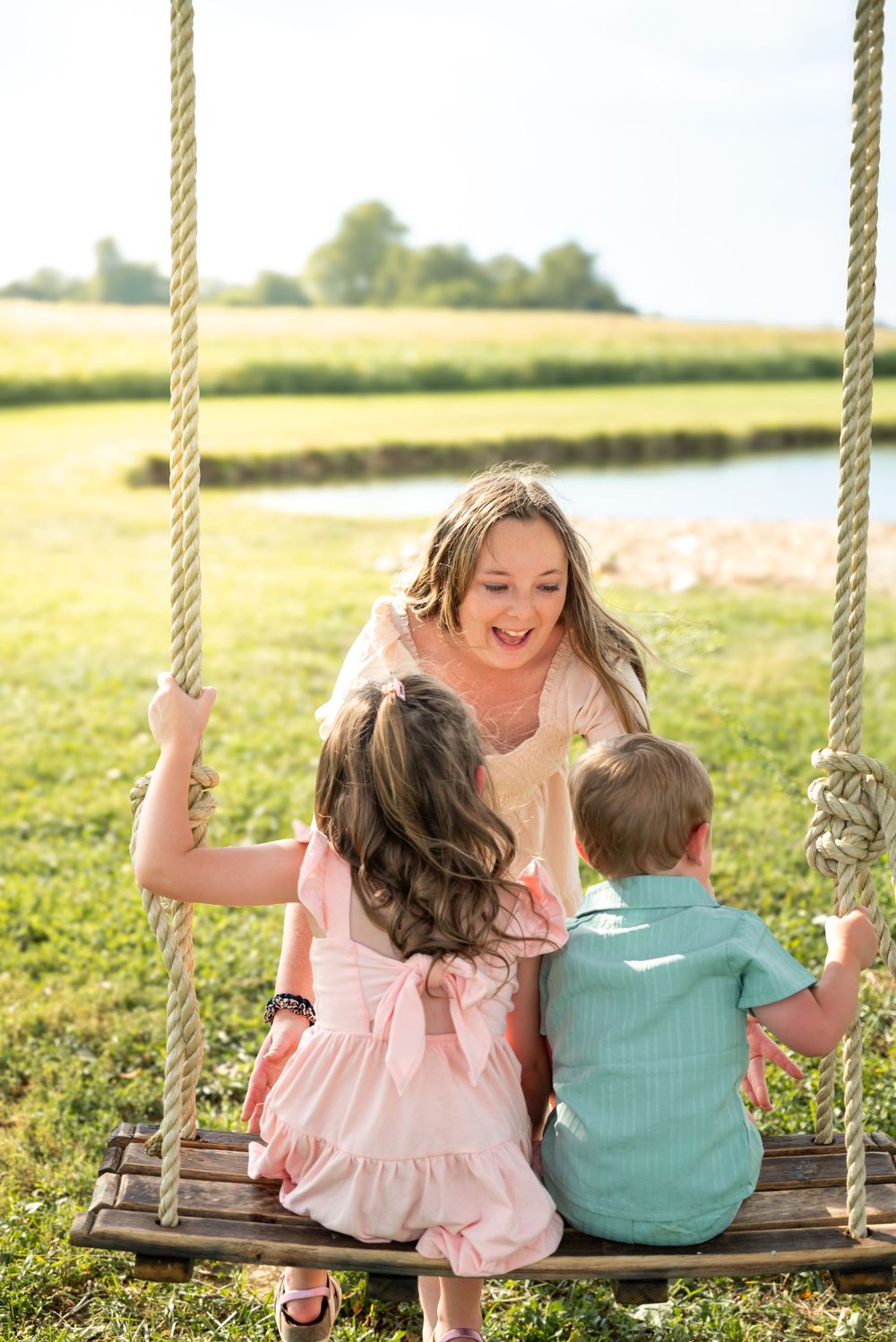 A mother sits on a wooden rope swing laughing with her two young children outdoors on a sunny day.