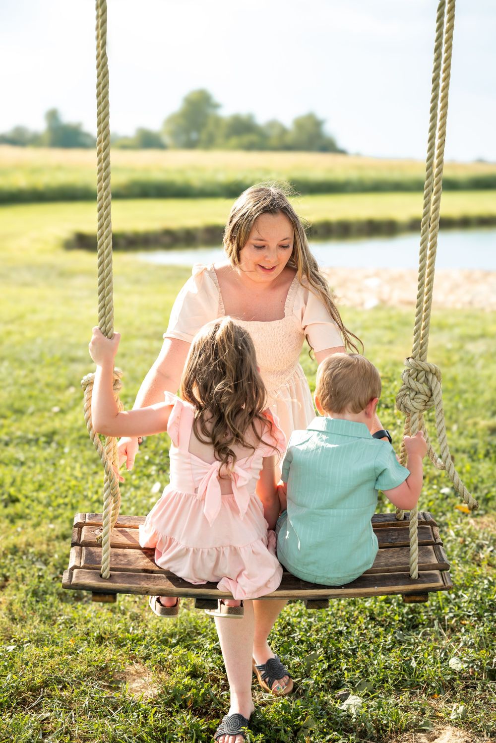 A mother in a white dress pushes two young children on a wooden rope swing in a sunny green field.
