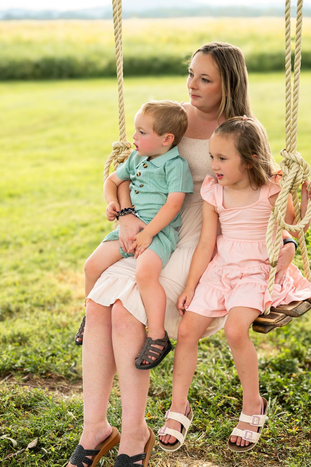 A mother sits on a rope swing with her two young children in a green field, wearing summer outfits.