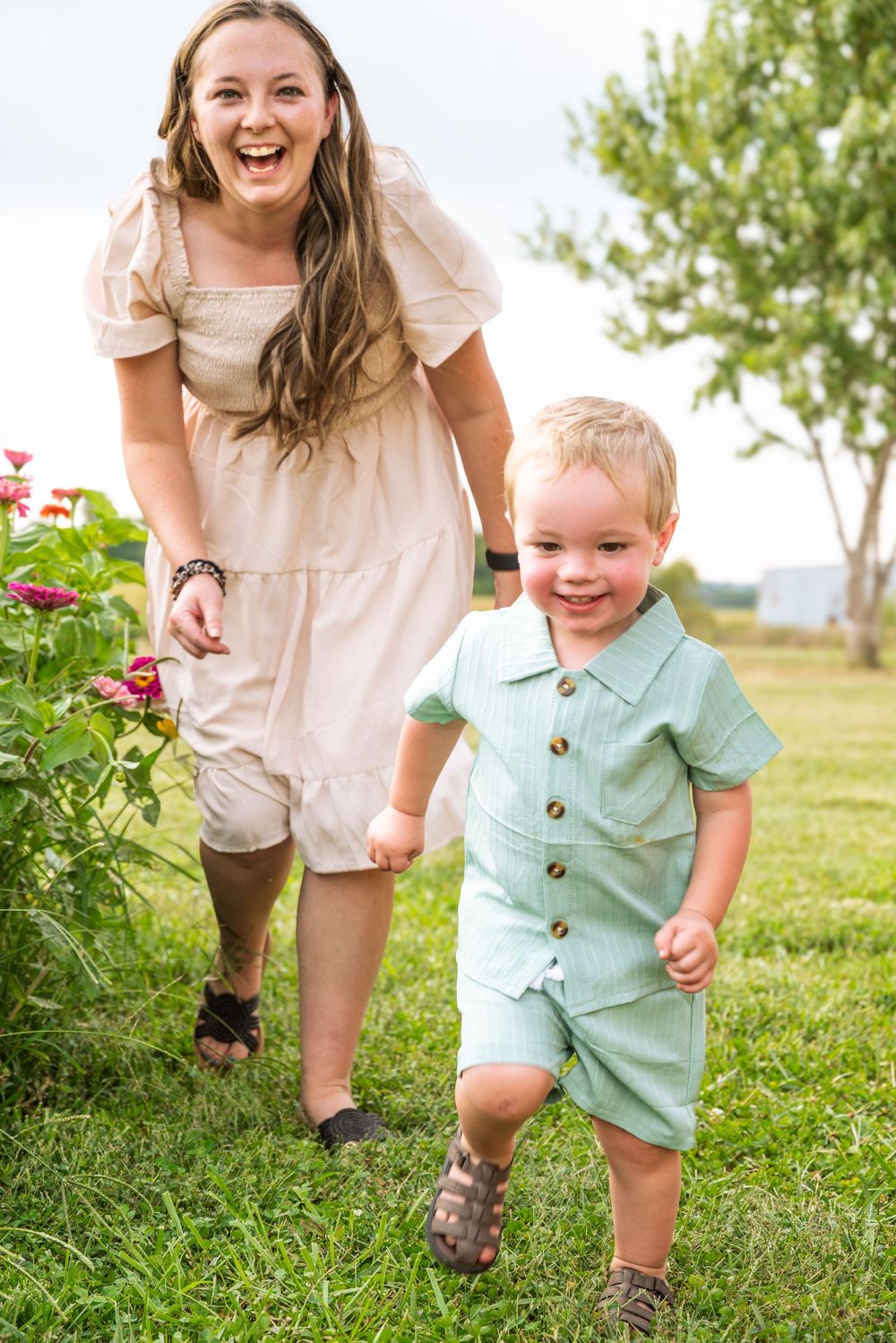 A laughing woman in a white dress holds hands with a smiling toddler boy in a mint romper running through green grass.