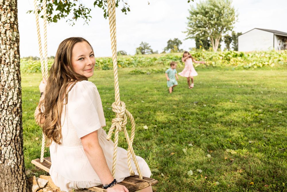 Young girl sits on a wooden rope swing by a tree, smiling, while two children play on a green lawn behind her.