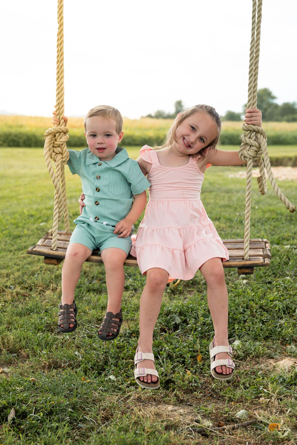Two young children sitting together on a rope swing outdoors, boy in mint outfit and girl in pink dress.
