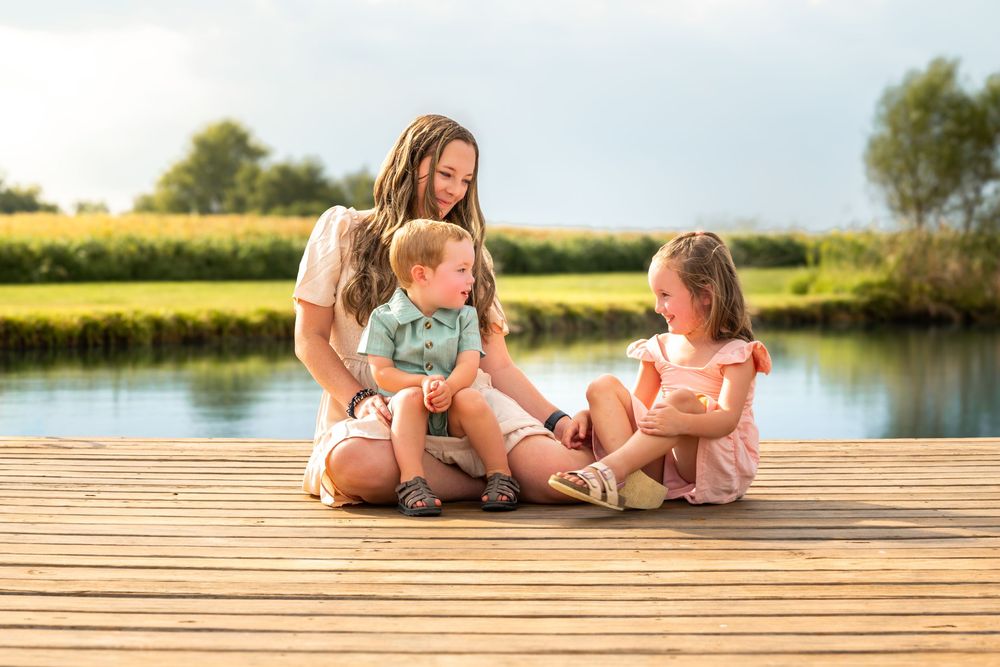 A mother sits on a wooden dock with two young children by a peaceful lake on a sunny day.