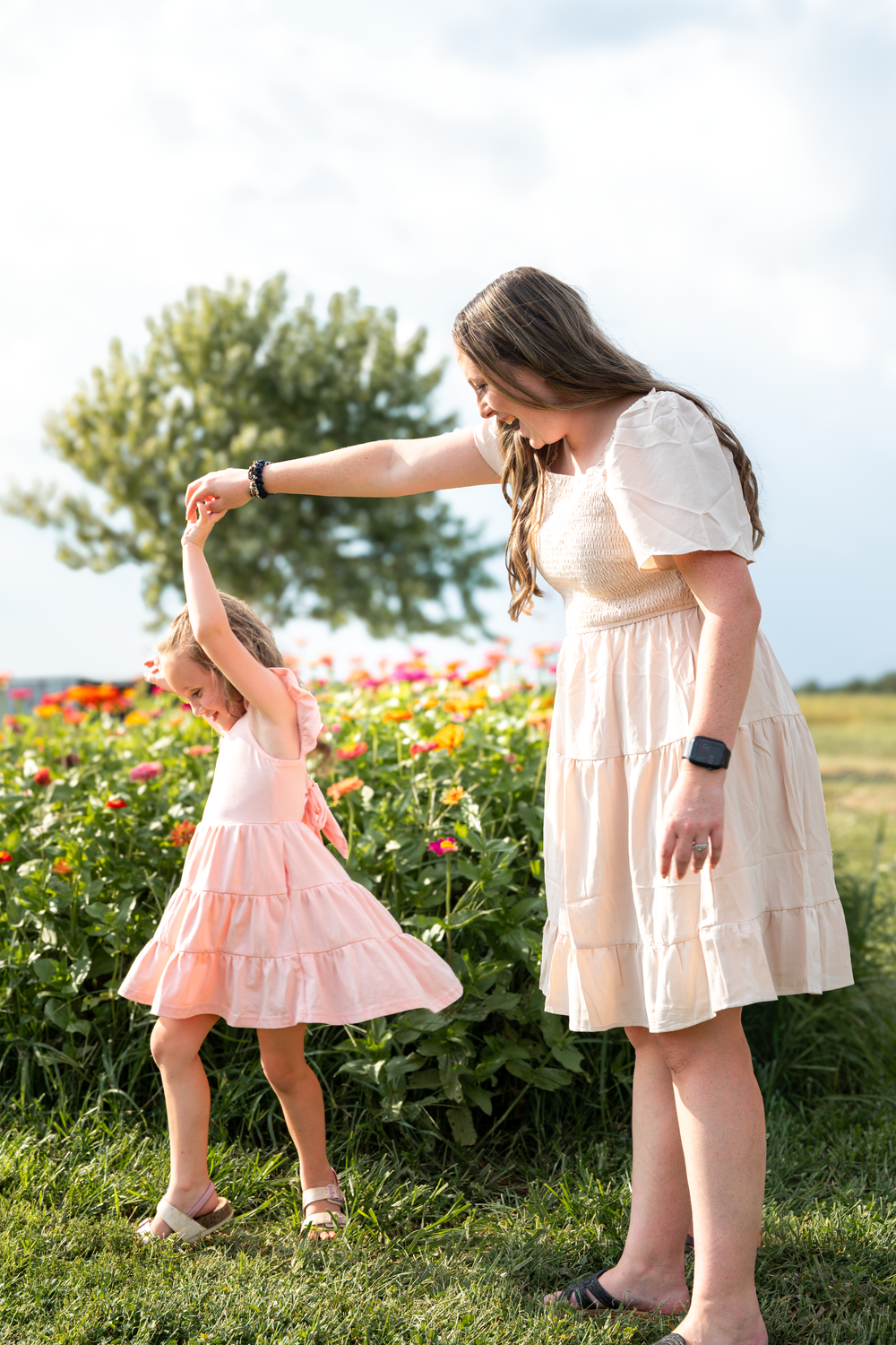 A mother twirls her young daughter in matching dresses outdoors near a colorful flower garden on a sunny day.