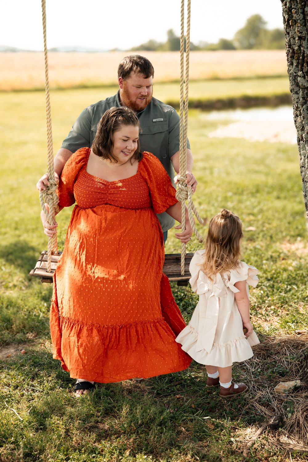 A family poses outdoors near a tree swing, with a pregnant woman in orange dress, bearded man, and young girl in white dress.