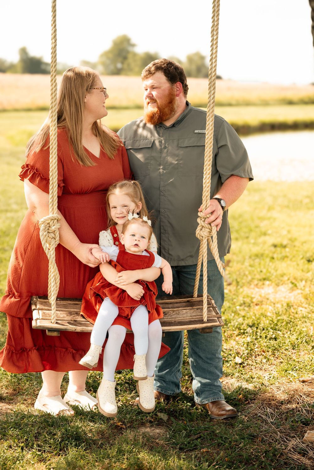 Family of four posing on a wooden rope swing outdoors, parents in red and green with two young daughters in matching red dresses.