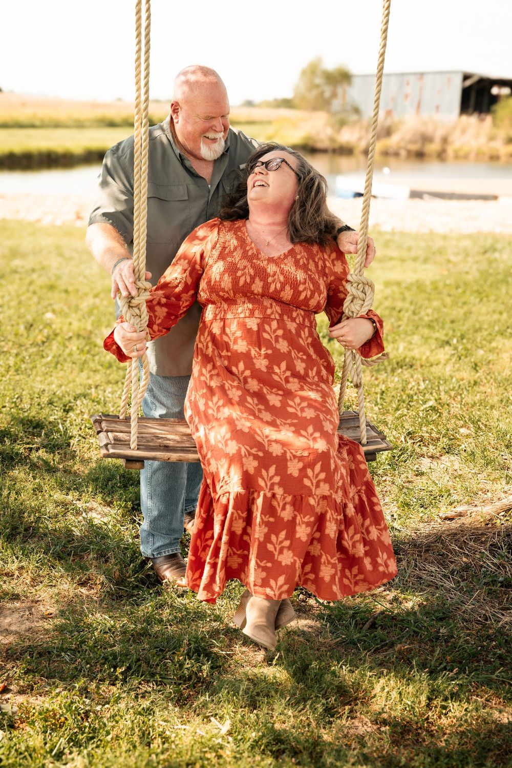 A joyful older couple sharing a playful moment on a wooden rope swing outdoors near a pond.