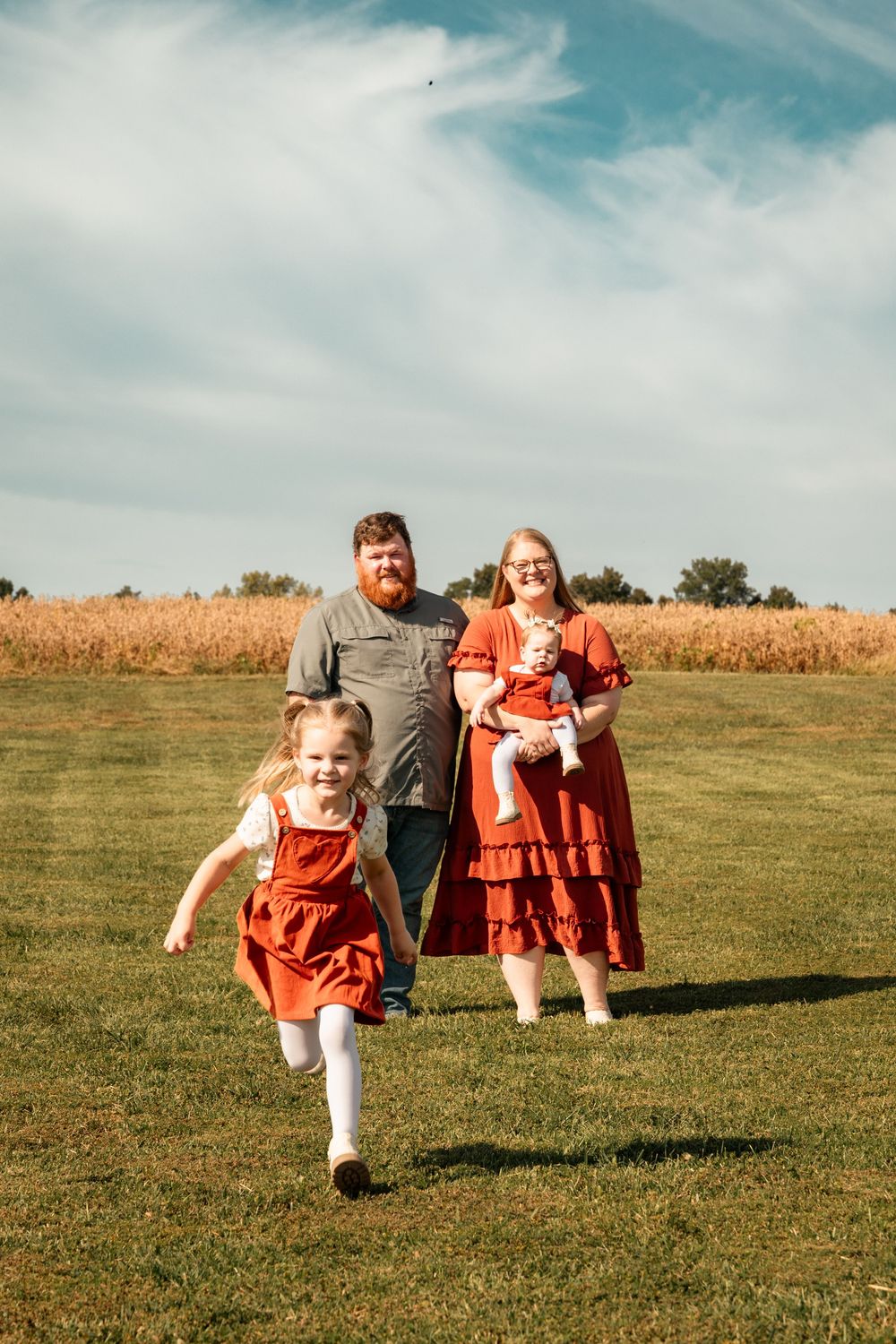Family of four enjoying a sunny day in a green field near a cornfield, with a girl in red running toward the camera.