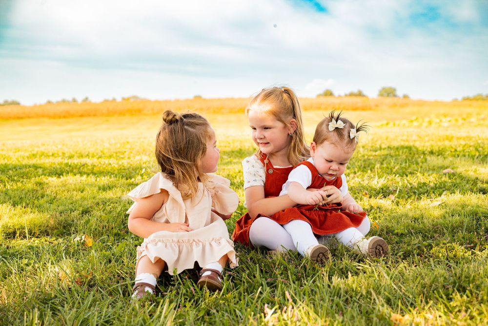 Three young girls sitting together on green grass in a sunny field, wearing colorful dresses and smiling happily.