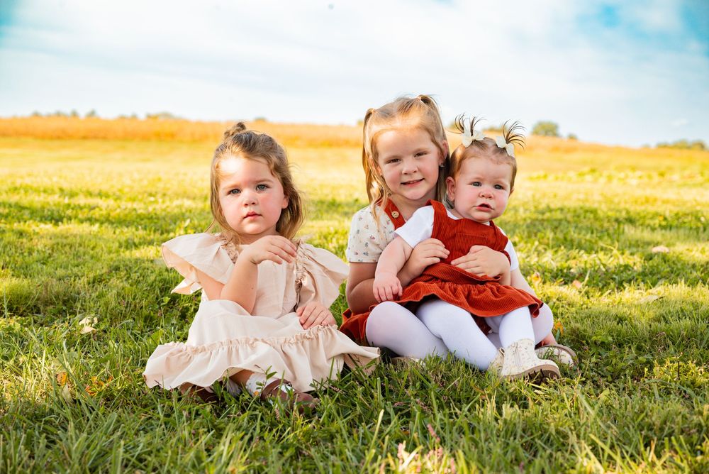 Three young girls in dresses sitting together on green grass in a sunny field, posing for an outdoor portrait photo.