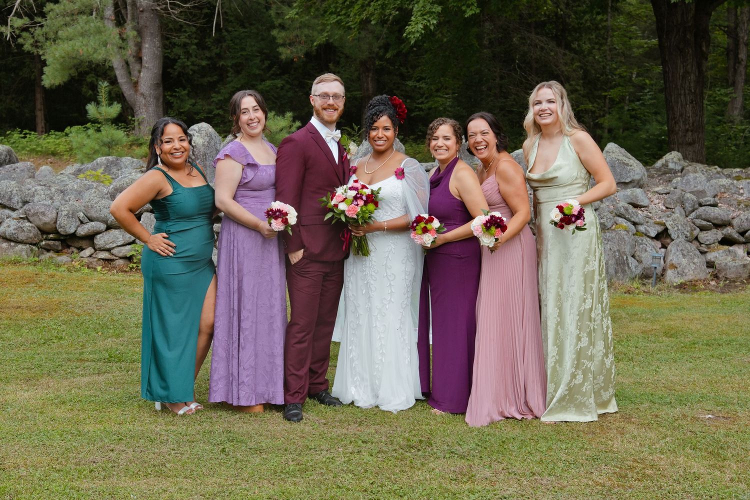 Wedding party poses outdoors near stone wall, groomsmen in tan suits, bridesmaids in purple and green gowns, bride and groom centered.