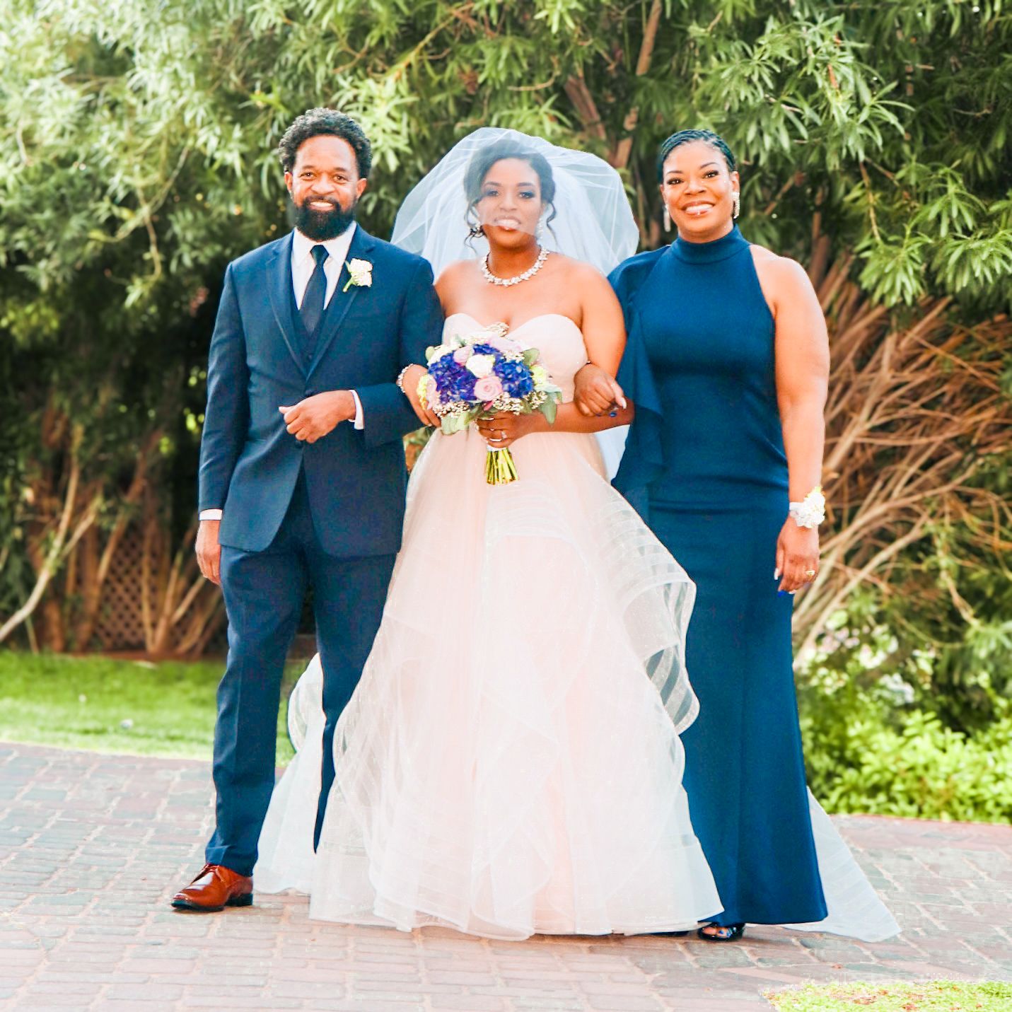 Bride in white gown holding blue bouquet stands with groom in navy suit and bridesmaid in blue dress outdoors.