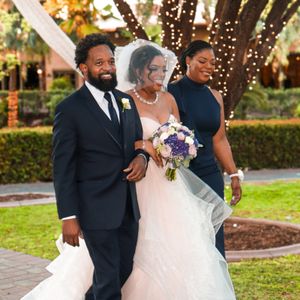 Bride in white gown with veil holds bouquet, escorted by two guests in navy attire at an outdoor wedding.