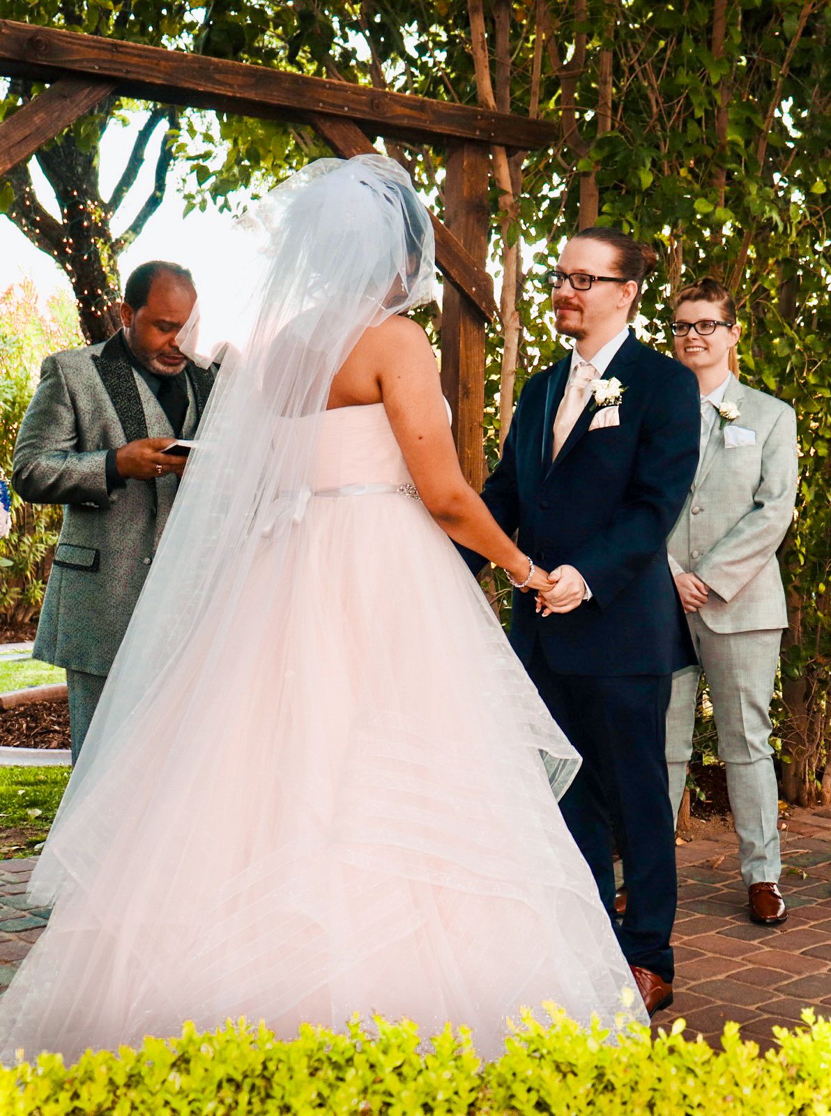 Outdoor wedding ceremony with bride in white gown and veil, groom in navy suit, officiant, and witness in gray suit.