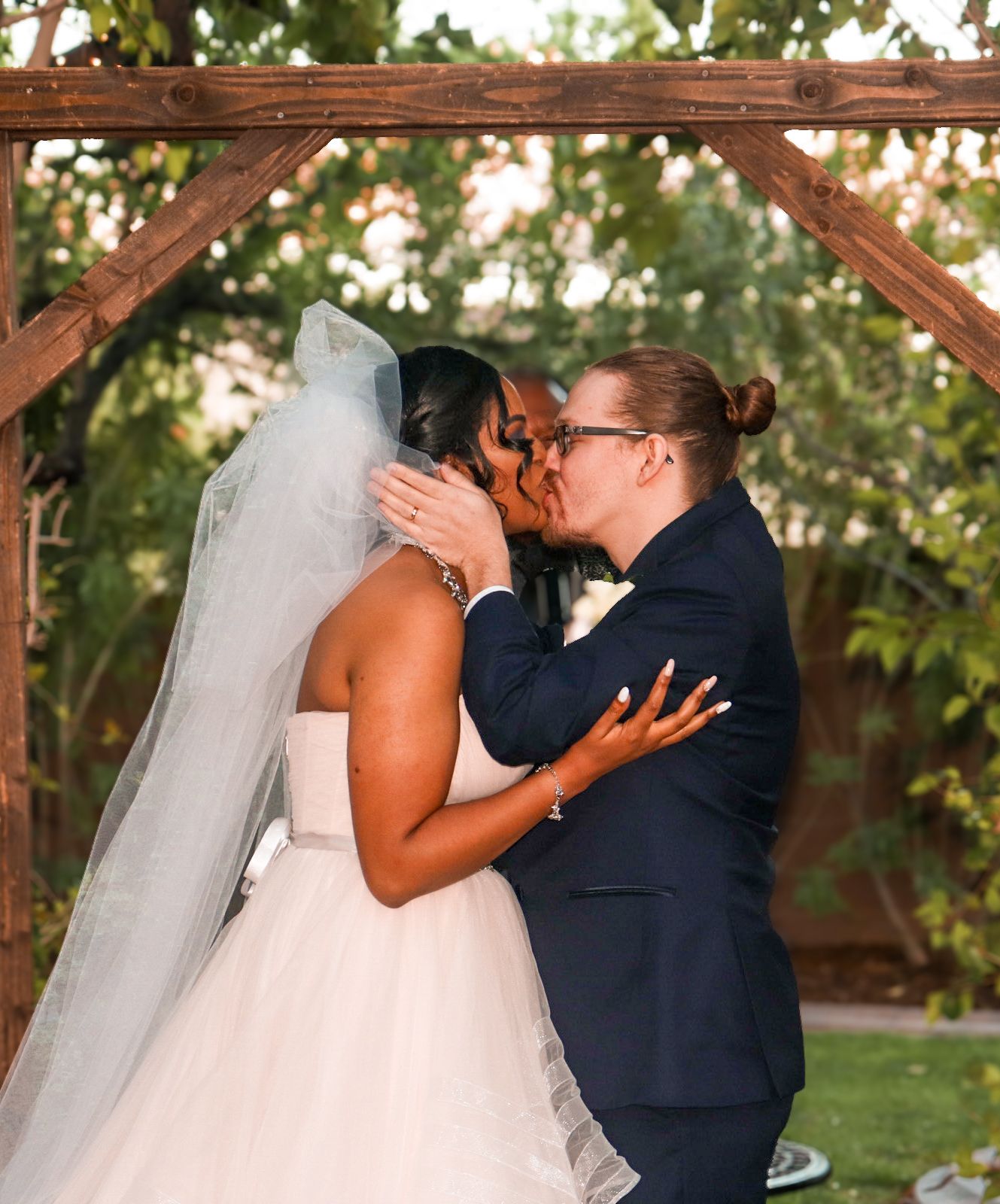 Two brides share their first kiss at an outdoor wedding ceremony under a rustic wooden arch.