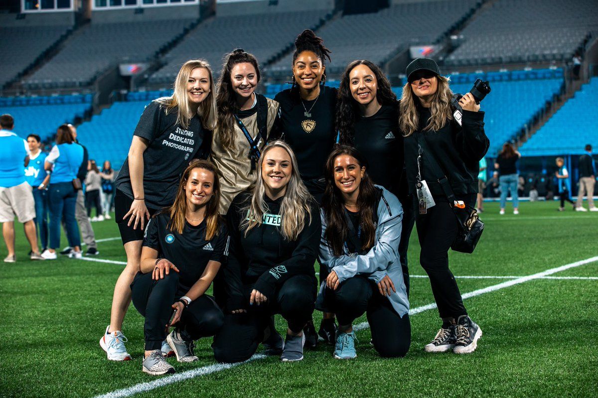 Group of nine women in black outfits posing together on a green stadium field during a sports event.