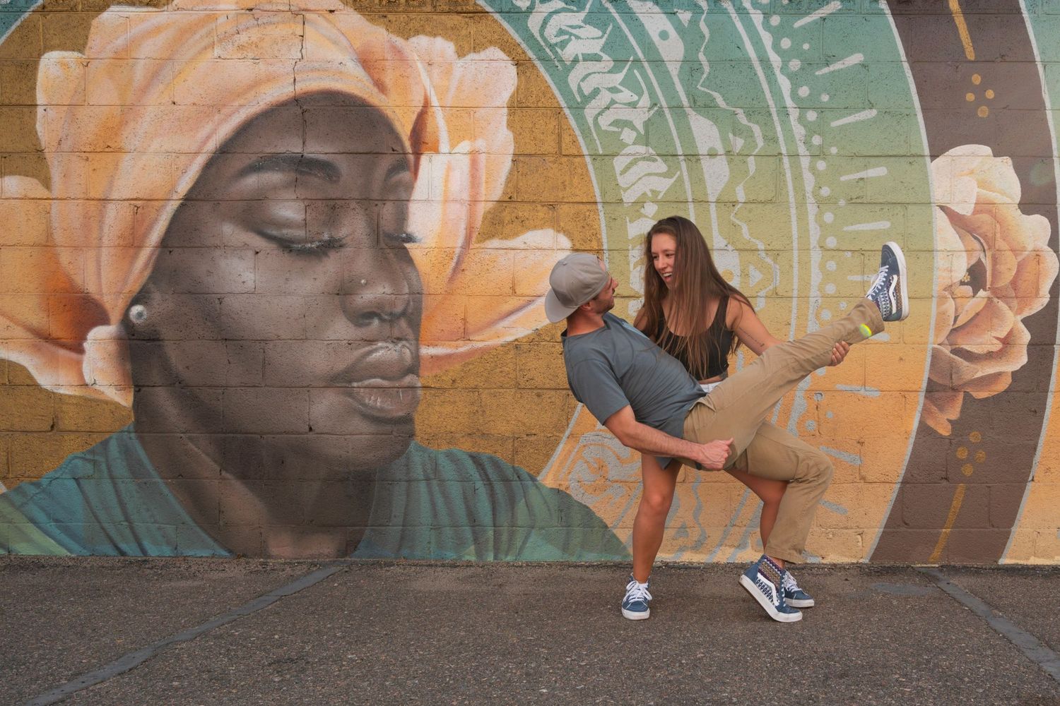 Couple dancing playfully in front of a colorful street mural featuring a woman with a floral headwrap.
