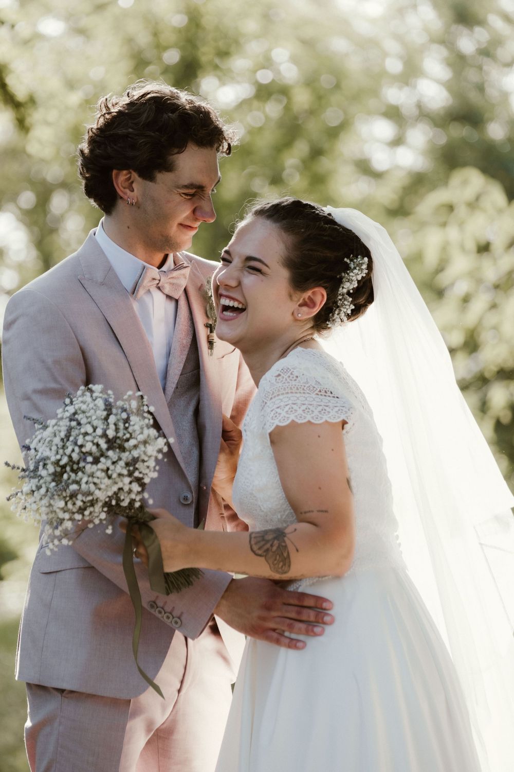 Laughing bride in white veil embraces groom in blush suit outdoors, holding baby's breath bouquet on wedding day.