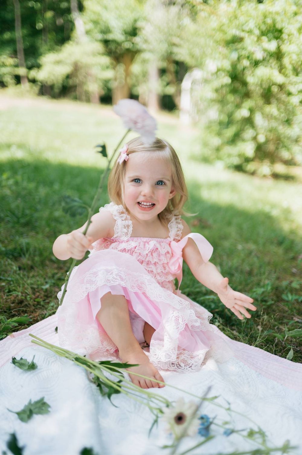 Happy blonde toddler girl in pink lace dress laughing and playing with flowers on a blanket in a garden in North Carolina