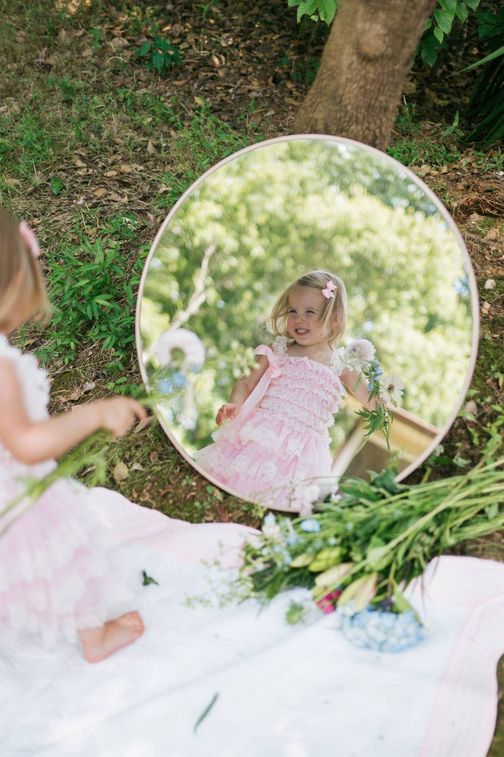Blonde toddler girl in pink ruffled dress looking at her reflection in a round mirror outdoors in a garden in North Carolina