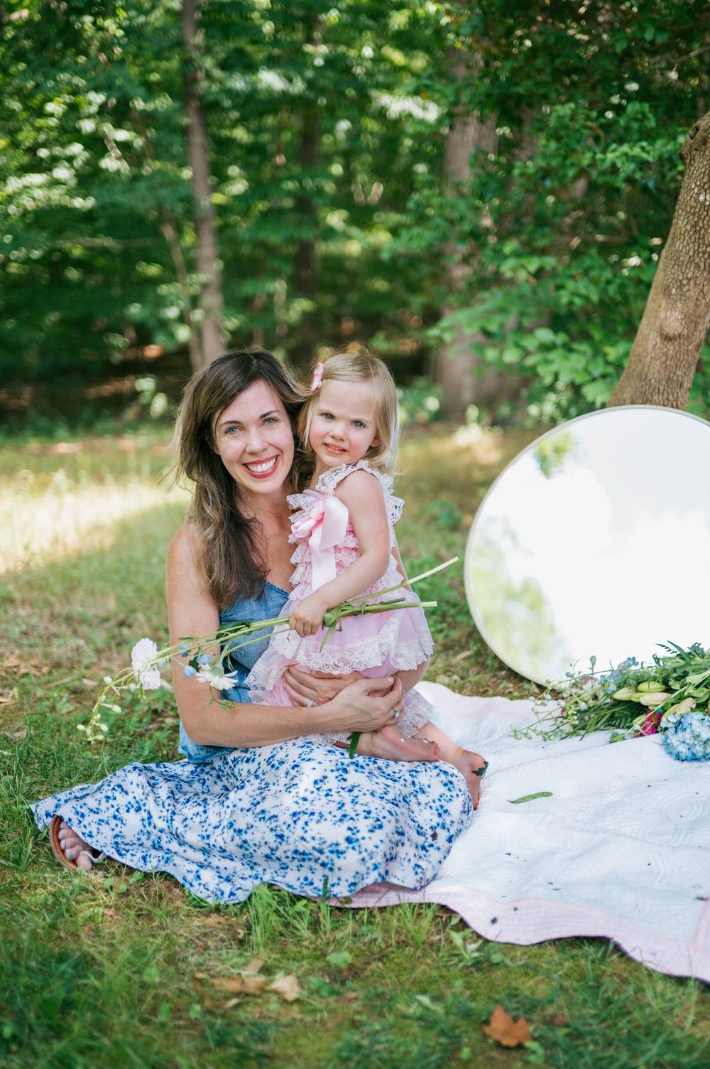 Laughing mother holding blonde toddler in pink lace dress on a blanket in a lush green outdoor setting in North Carolina