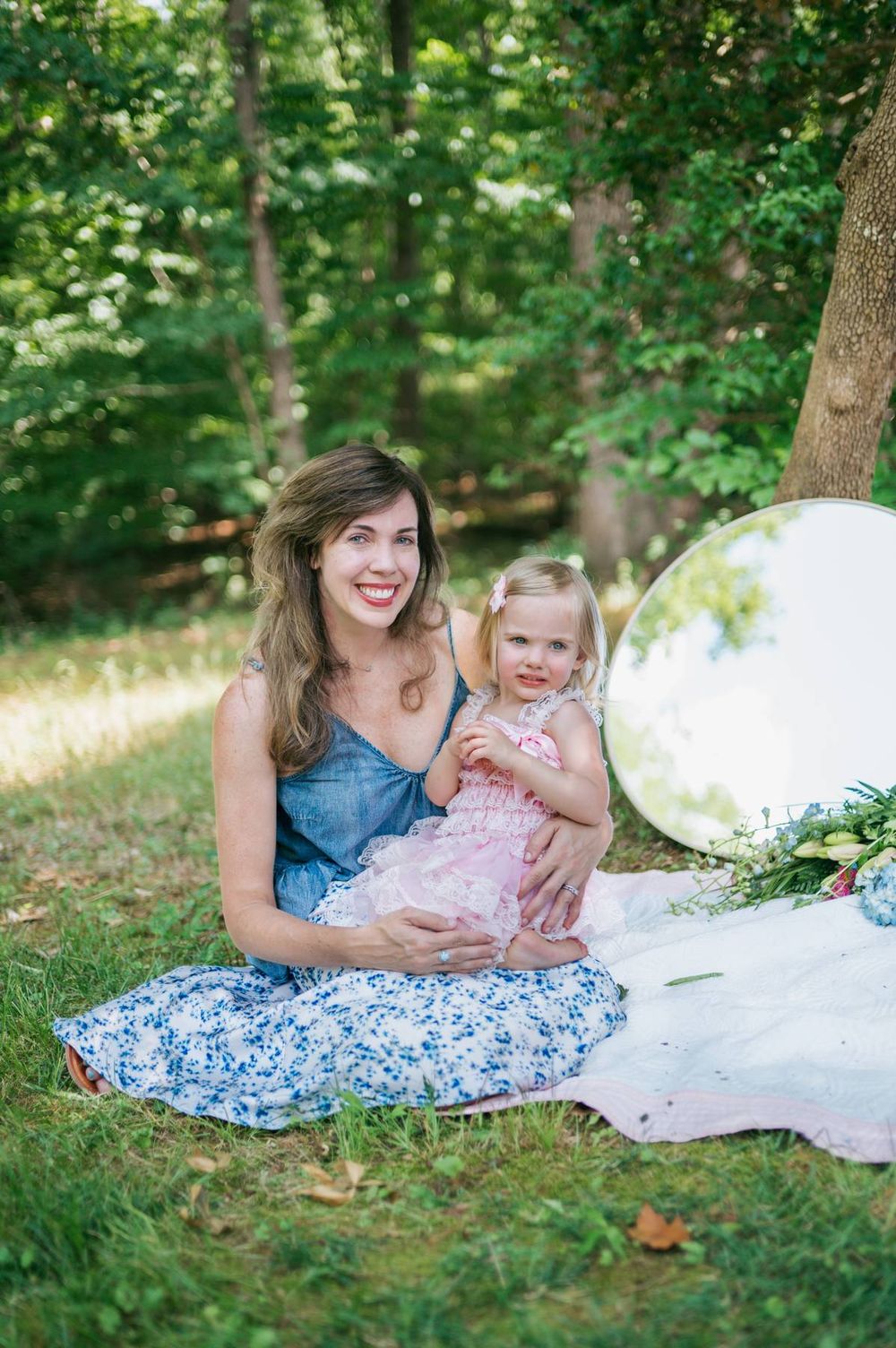 Smiling mother in blue floral dress sitting on grass holding her toddler daughter in a pink lace dress in North Carolina