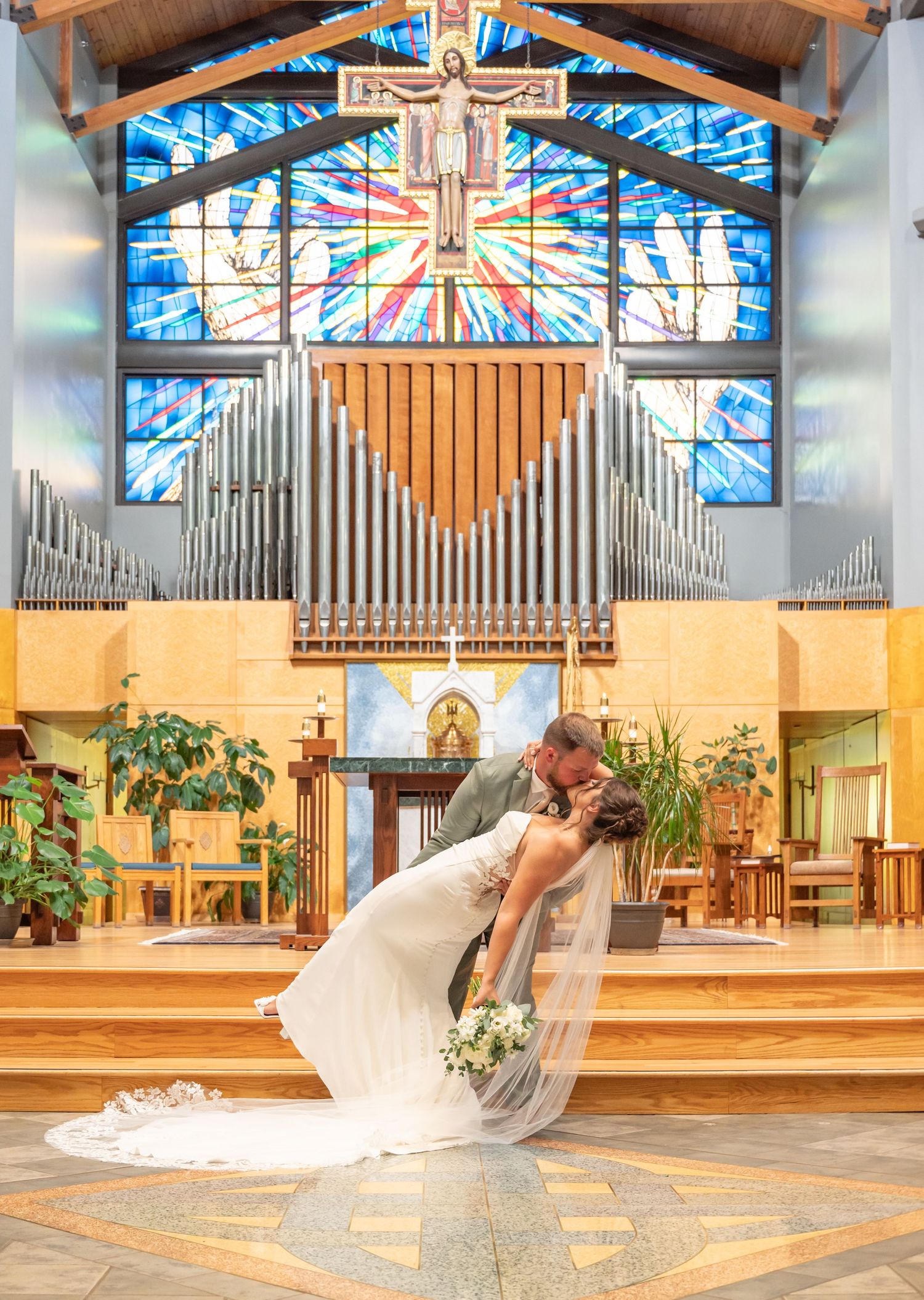 Bride and groom share romantic dip kiss before stunning stained glass window in church.