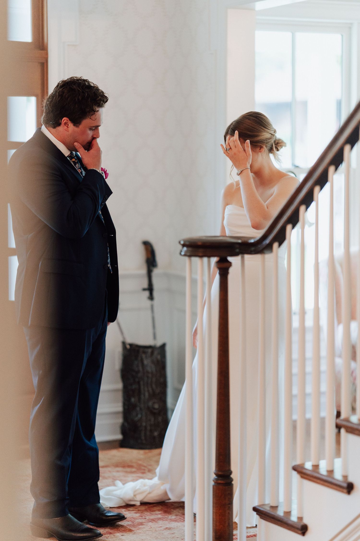 Emotional bride and groom share a first look moment on a staircase, both covering their faces with tears of joy.