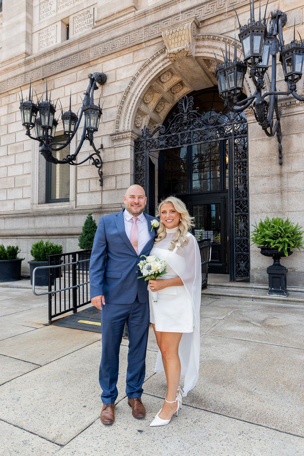 Bride in white mini dress and groom in navy suit pose smiling outside an elegant building entrance with ornate ironwork.
