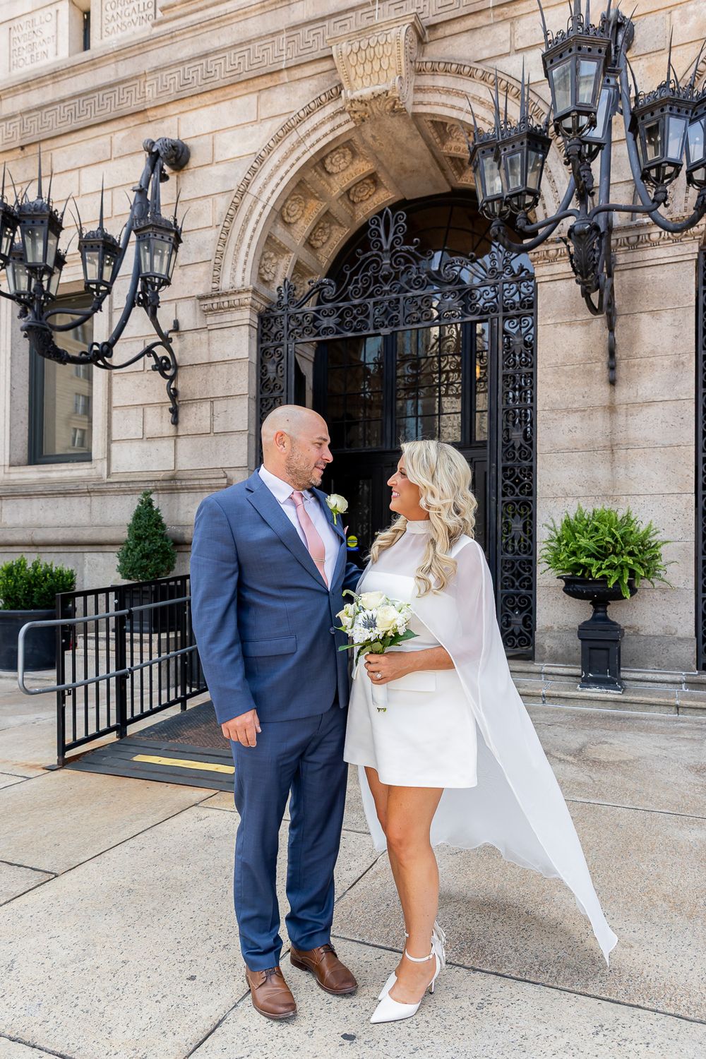 Couple in wedding attire poses outside ornate building, bride in white mini dress with veil, groom in blue suit.