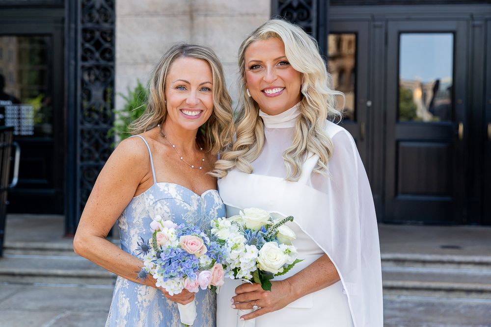 Bride in white gown holding blue and white bouquet poses with bridesmaid in blue floral dress outside elegant venue.