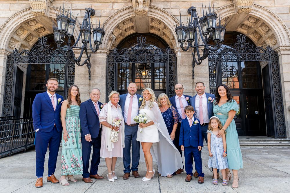 Wedding party poses in front of ornate stone building with iron gates, bride in white dress surrounded by family.