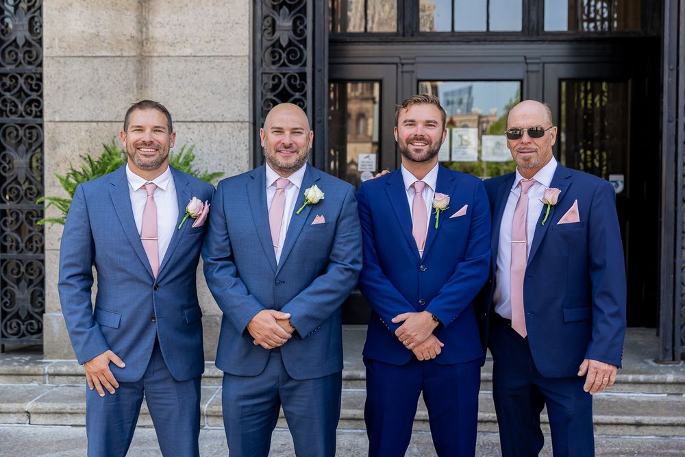 Four groomsmen in blue suits with pink ties and boutonnieres posing together outside a wedding venue.