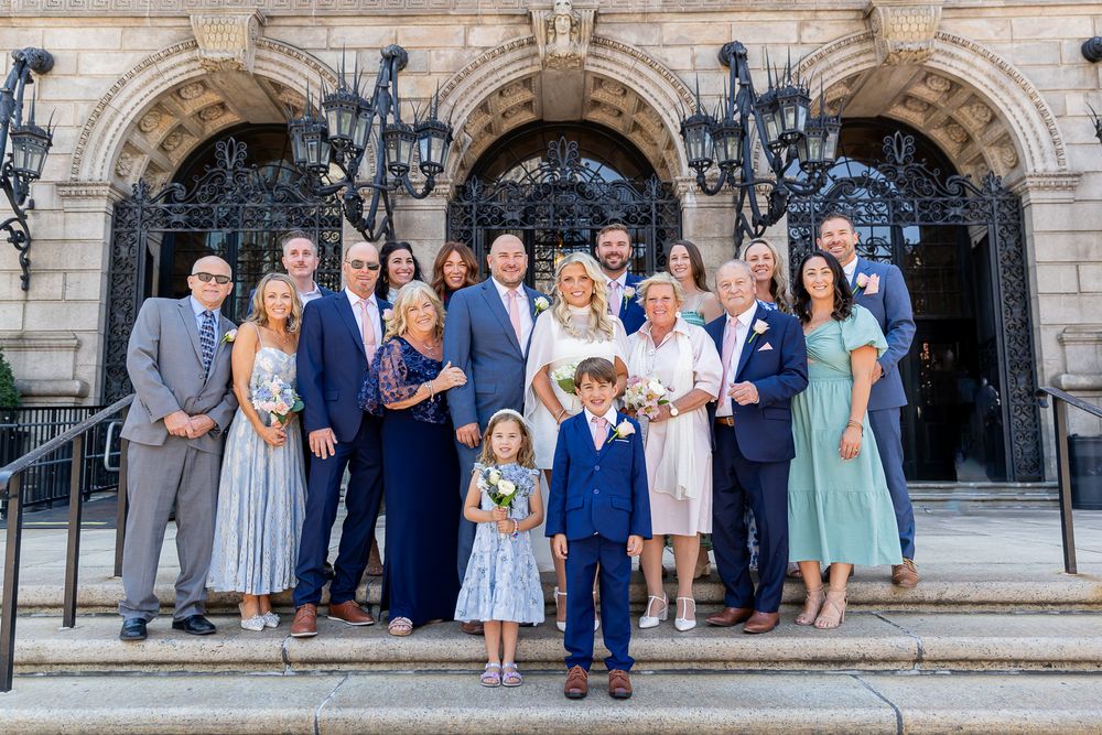A wedding party group photo posed on steps of an elegant historic building in formal attire.