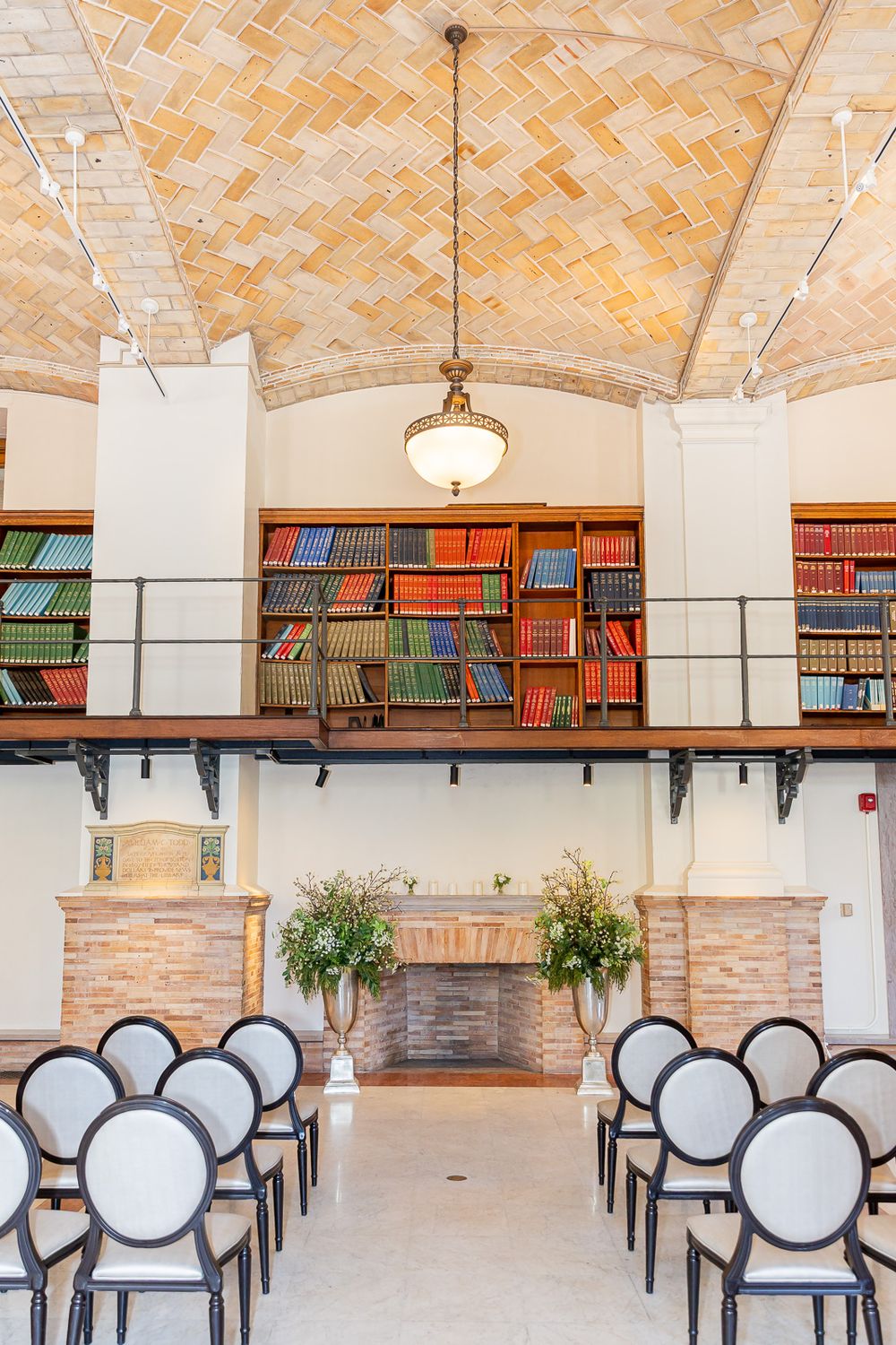 Elegant library venue with brick vaulted ceiling, colorful bookshelves, and white chairs arranged for a wedding ceremony.