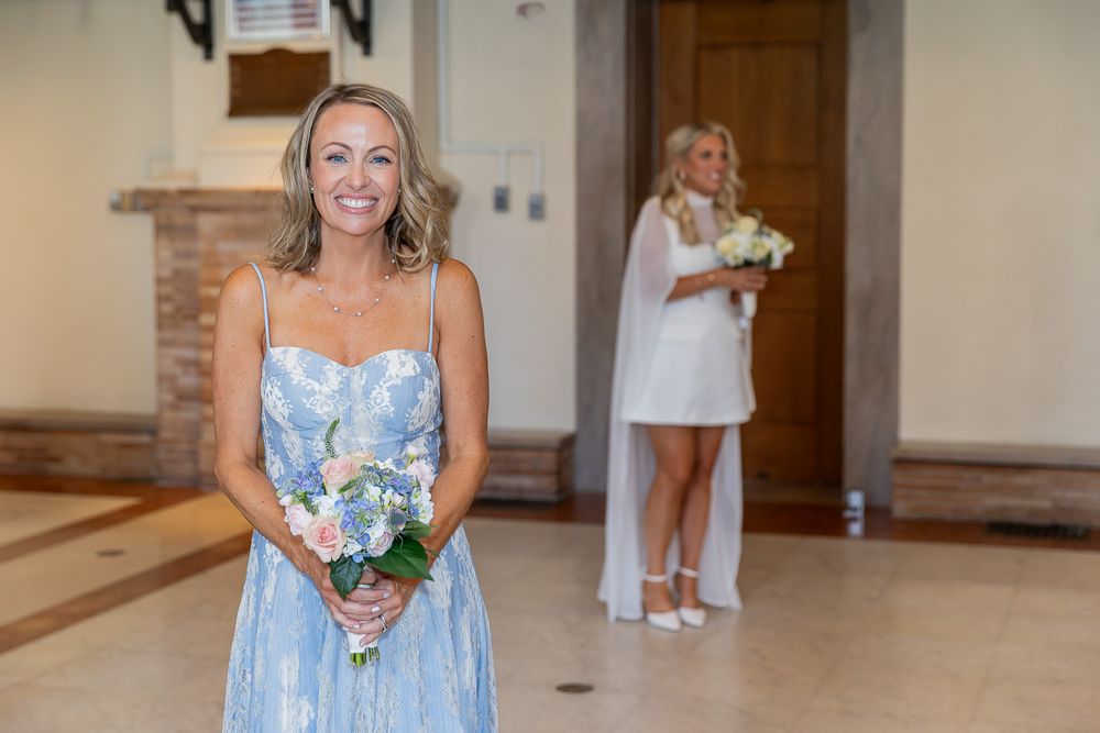Bridesmaid in blue floral dress holds bouquet indoors, with bride in white dress visible in background.