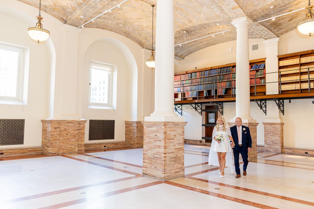Bride and groom walking through elegant wedding venue with tall arched ceilings, columns, and library bookshelf backdrop.