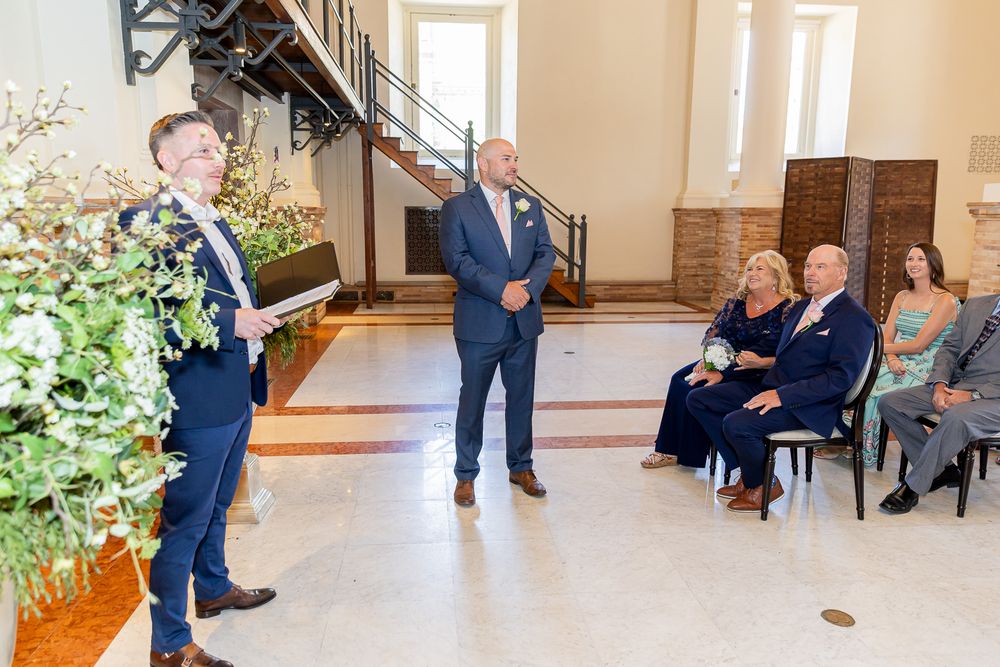 Groom stands at wedding altar in navy suit while guests seated in elegant hall watch the ceremony.