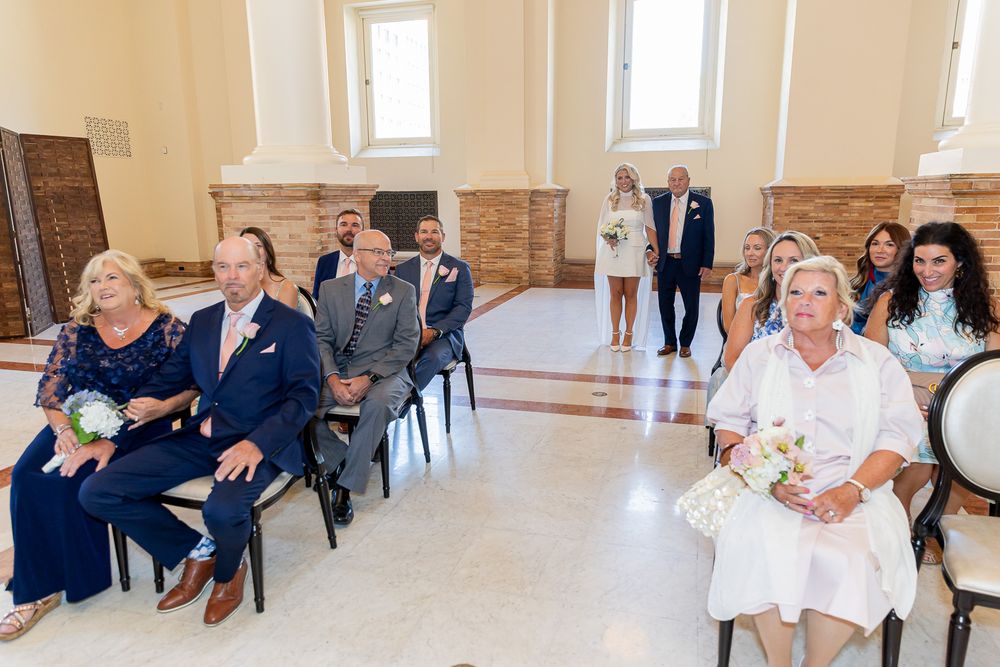 Wedding guests seated in elegant venue watching ceremony, women holding bouquets in foreground, couple visible at altar.