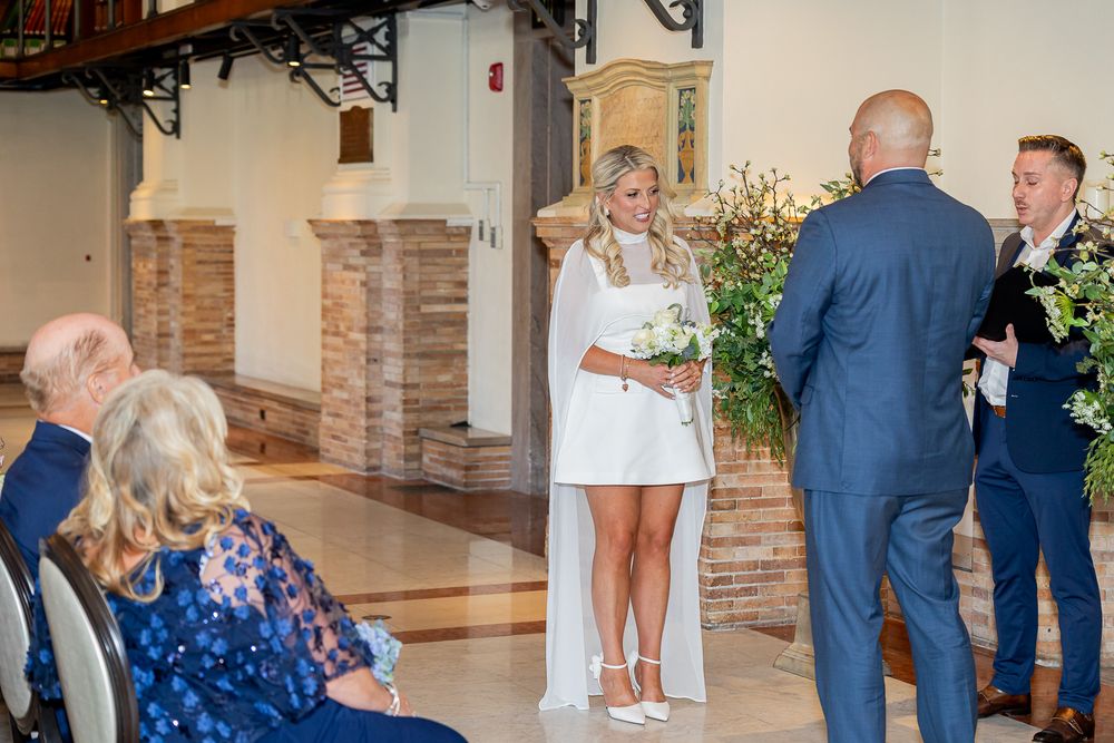 Bride in white mini dress holding bouquet stands at wedding ceremony altar with groom and officiant in elegant venue.
